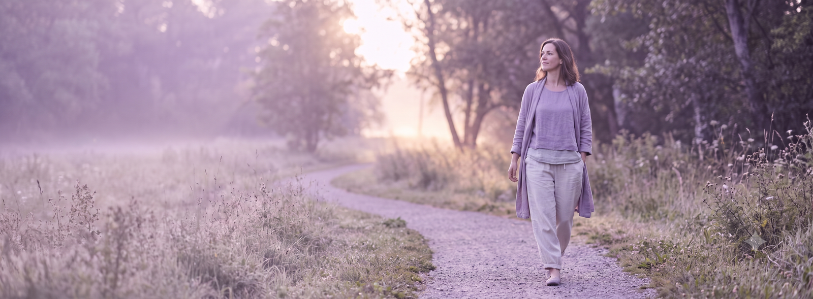 Woman walks along a winding gravel path through a misty purple-hued meadow at sunrise, wearing light lavender clothing.