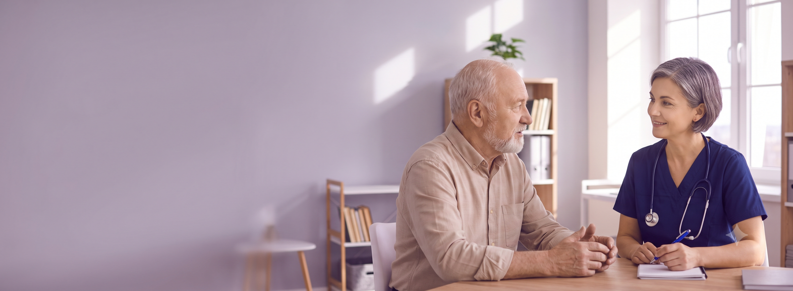 Nurse or doctor in navy scrubs with stethoscope talking with an elderly man at a wooden table in a bright clinic setting.