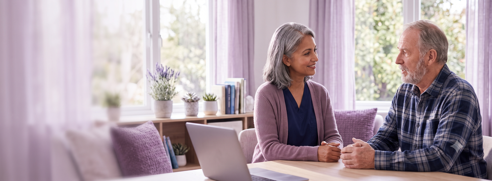 Older woman and man sit facing each other at a wooden table, smiling and talking in a bright living room with purple curtains and a laptop nearby.