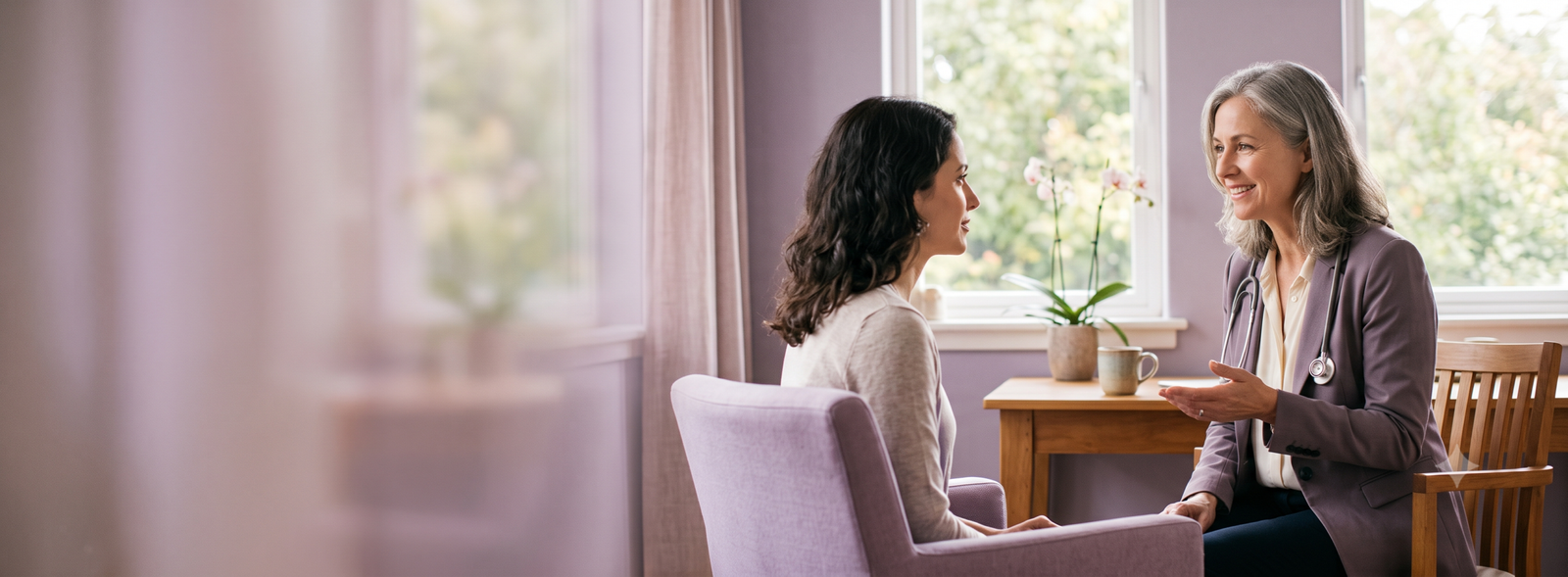 Female clinician with a stethoscope talks with a patient in a purple themed consultation room