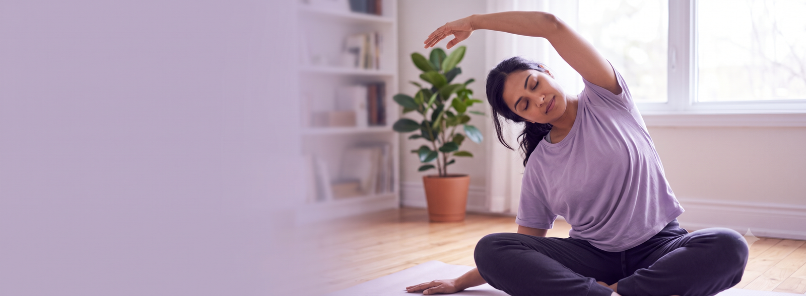 Woman in a purple shirt performing a seated side stretch on a yoga mat in a bright living room