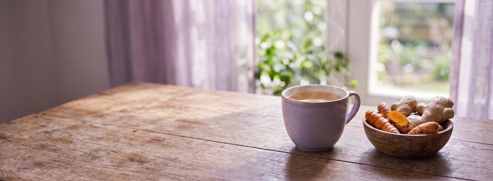 Purple mug of steaming tea on a rustic wooden table beside a bowl of cookies near a sunlit window with lavender curtains.