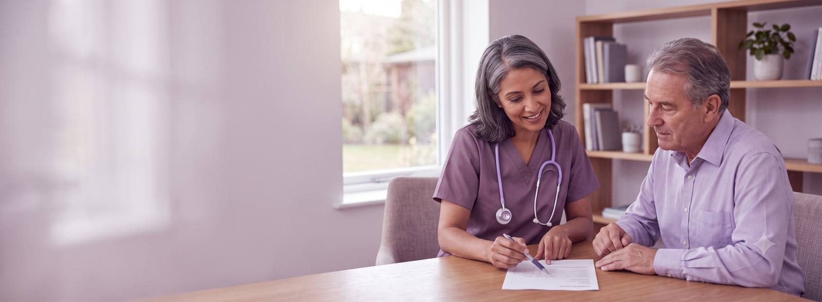 Nurse with stethoscope sits with an elderly man at a table, reviewing a form together.