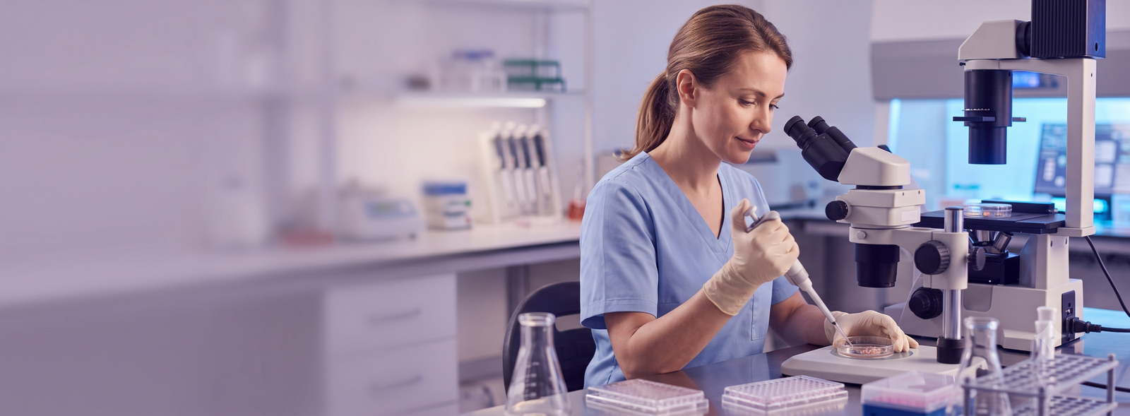 Female scientist in blue scrubs using a micropipette at a microscope in a modern lab