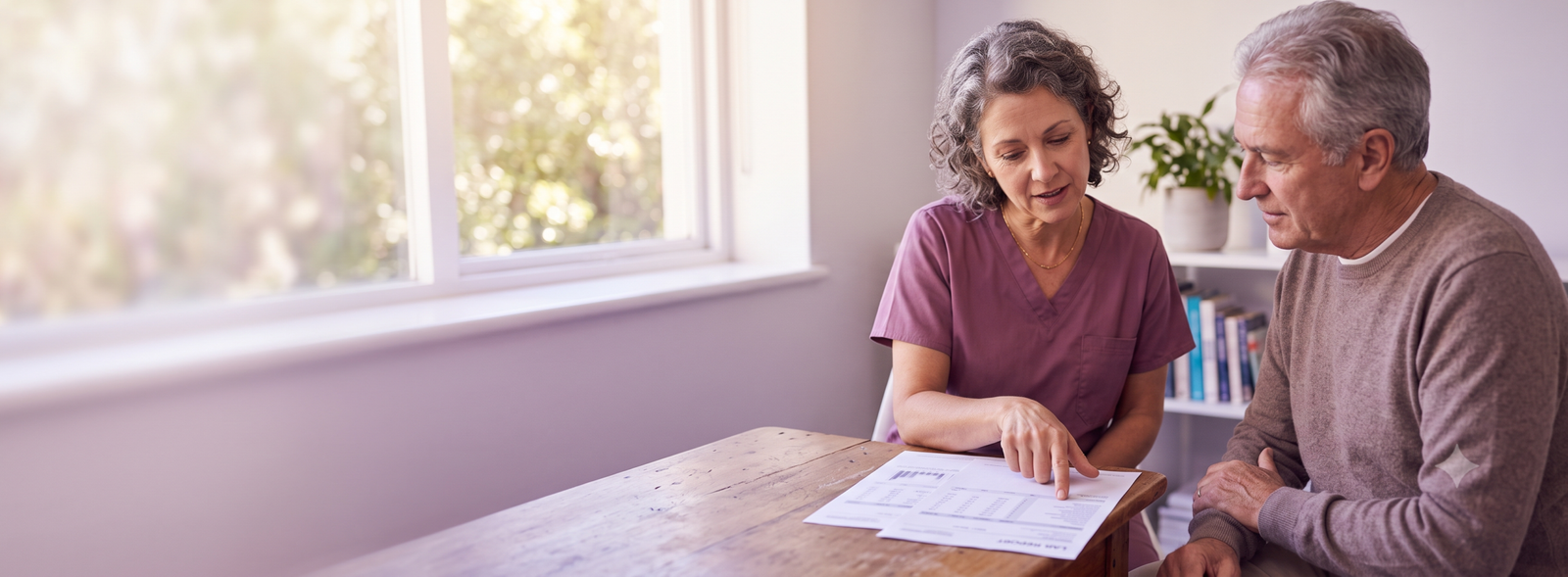 GeminiGeneratedImagex024zox024zox024 Samarth IVF Caregiver in purple scrubs explains a document to an elderly man at a wooden table in a bright room by a window a paperwork consultation scene at home