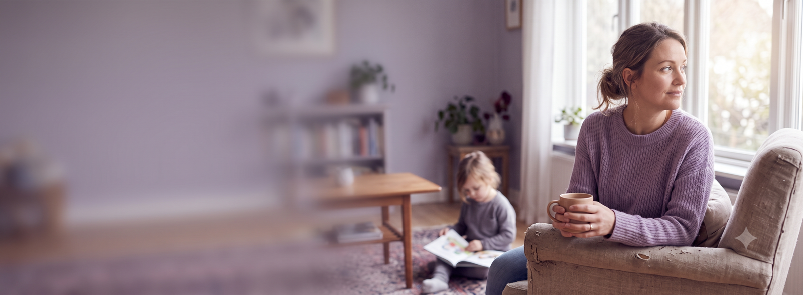 Woman in a lavender sweater sits on a beige sofa by a window, holding a mug as a child reads a book on the floor nearby in a cozy living room.